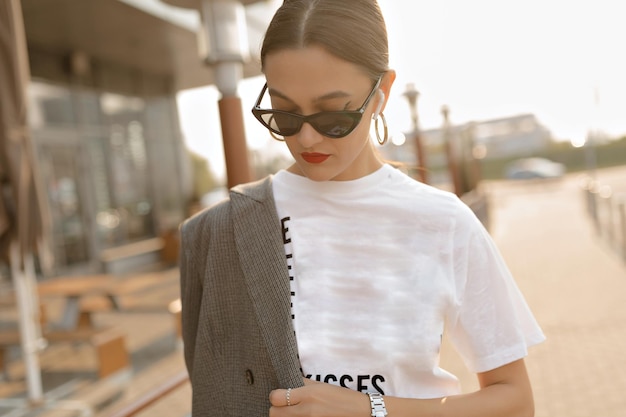 Portrait os stilvolle Frau mit Sonnenbrille, die auf der Straße läuft. Sie trägt ein weißes T-Shirt, hat rote Lippen und schaut nach unten