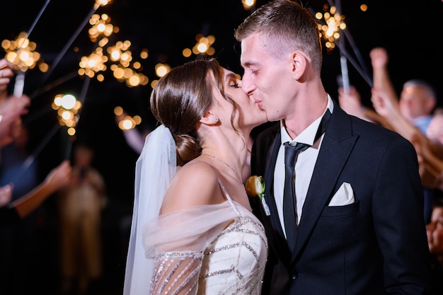 Portrait von Liebespaar küssen feiern Hochzeitstag mit Gästen Freunde im Hintergrund Night Party der Bräute Schöne Brünette Frau im eleganten Hochzeitskleid küssen Bräutigam Sparkles Happy