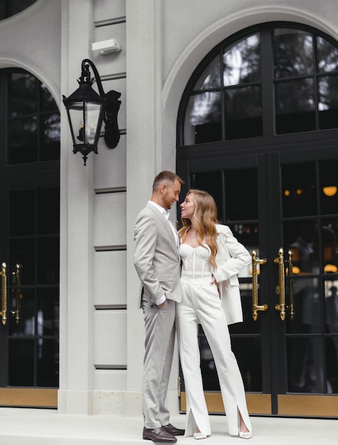 Portrait von stilvollen Bräuten Paar Mann und Frau im weißen Hochzeitsanzug, die einander betrachten Posing in der Nähe eines modernen Gebäuderestaurants Moderne Hochzeit Glückliches Ehepaar Ehe