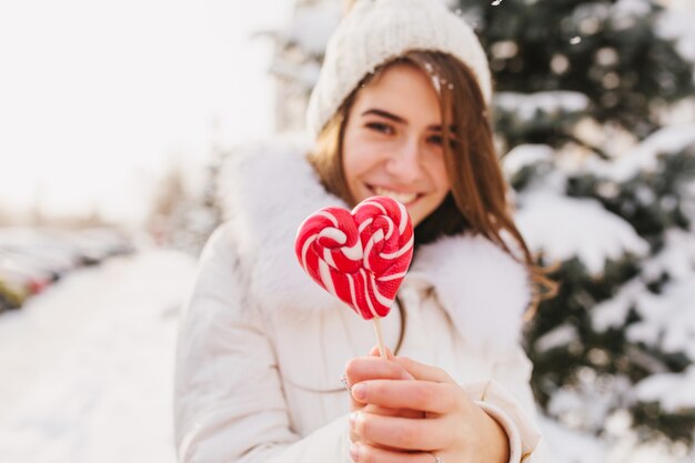 Portrait Winter junge Frau hält rosa Herz Lollypop, Chillen auf Straße voll mit Schnee in sonnigen Morgen. Weiße Strickmütze, lächelnd.