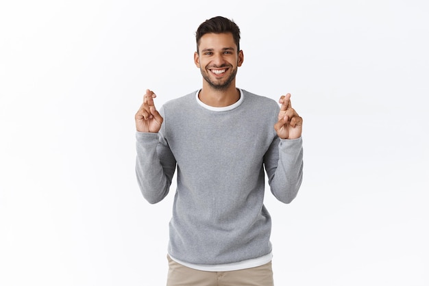 Positiv denkendes gebets- und glaubenskonzept hoffnungsvoller lächelnder optimistischer gutaussehender kerl in grauem pullover kreuzt die finger für viel glück, erwartet gute nachrichten und wartet auf den weißen hintergrund eines netten deals