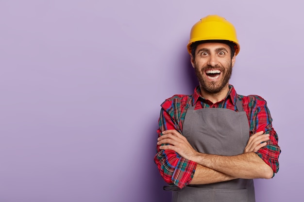 Kostenloses Foto positiv glücklicher vorarbeiter in arbeitskleidung, verschränkt die arme, trägt gelben helm