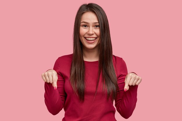 Positiv lächelnde Dame mit langen dunklen Haarspitzen mit beiden Vorderfingern nach unten, in Hochstimmung, empfiehlt, auf den Boden zu schauen