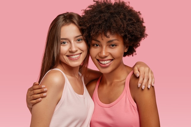 Positiv lächelnde verschiedene beste Freunde umarmen sich, haben freundschaftliche Beziehungen, posieren für ein gemeinsames Foto, freuen sich zu treffen, isoliert über rosa Wand. Interracial Freundschaft, Unterstützungskonzept