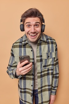 Positiv optimistischer mann hört lieblings-audiotrack in kopfhörern hält handy reagiert auf etwas lustiges, gekleidet in lässig kariertem hemd einzeln auf beigem hintergrund. emotionen konzept