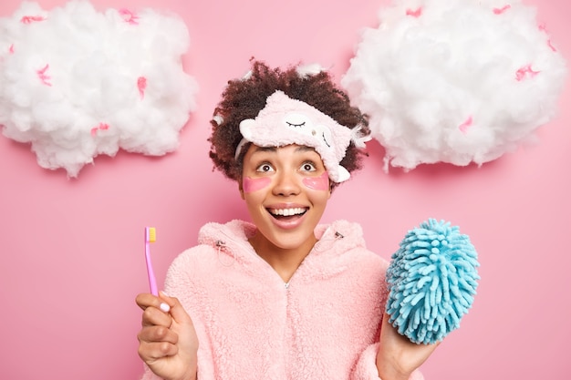 Positiv überrascht Afro-Amerikanerin konzentriert über trägt Schlafmaske und Pyjama hält Zahnbürste, um Zähne zu putzen trägt Schönheit Kollagen Flecken unter den Augen Posen zu Hause