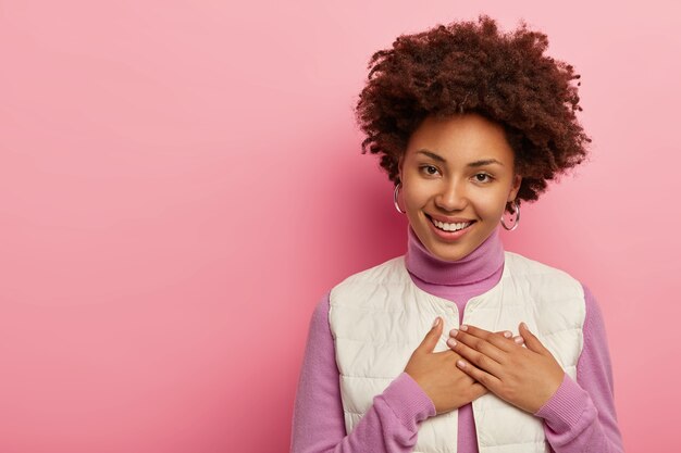 Positive Afro-Frau drückt Hände auf die Brust, schätzt herzerwärmende Worte, lächelt sanft isoliert über rosa Wand