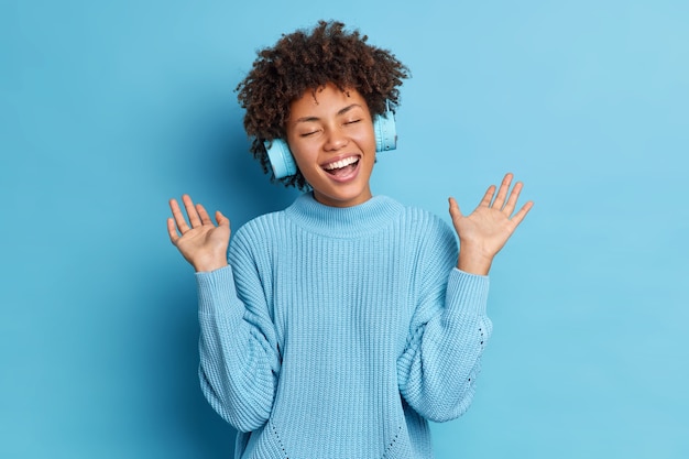 Positive Afroamerikanerin mit lockigem Haar hebt die Handflächen und hat Spaß, während das Hören von Audiospuren drahtlose Kopfhörer trägt, die in einen lässigen Pullover gekleidet sind