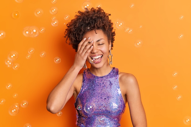 Positive afroamerikanische Frau bedeckt Gesicht mit Handlächeln hat im Großen und Ganzen perfekte weiße Zähne in stilvoller Kleidung, die über orangefarbener Wand mit Seifenblasen herum isoliert ist