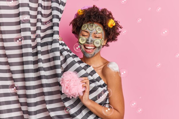 Positive afroamerikanische Frau singt Lied während des Duschens wendet Gurkengesichtsmaske an, um Haut zu verjüngen hält Schwamm genießt Körperpflegeverfahren isoliert auf rosa Wandseifenblasen herumfliegen