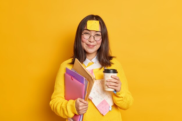 Positive asiatische Arbeiterin hält Einweg Tasse Kaffee hält Ordner hat Aufkleber mit Grafik auf der Stirn klebt hat Pause nach Prüfung Lernen.