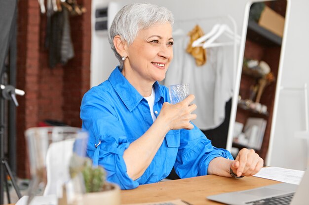 Positive attraktive reife Freiberuflerin, die neue gesunde Gewohnheiten entwickelt, vor einem offenen tragbaren Computer sitzt, ein Glas Wasser hält, sich während einer kleinen Pause erfrischt und freudig lächelt