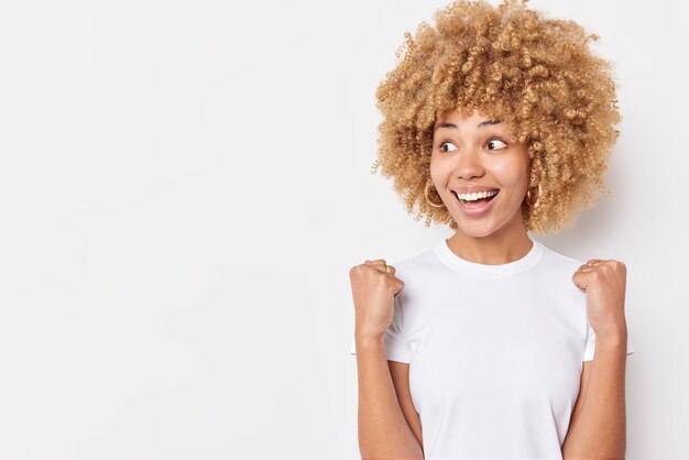 Positive aufgeregte lockige Frau macht siegreiche Geste ballt Fäuste feiert persönliche Erfolge konzentriert weg trägt lässiges Basic-T-Shirt isoliert über weißem Hintergrund Kopierraum für Text