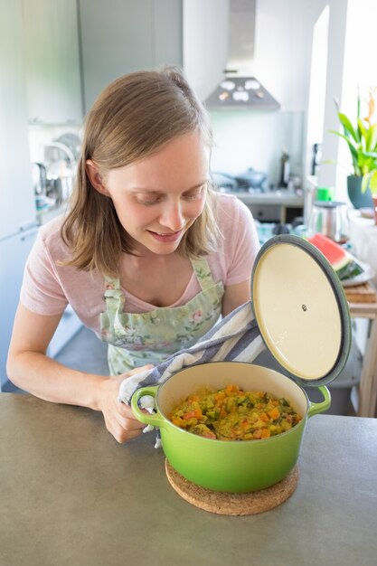 Positive Bloggerin, die in ihrer Küche kocht, Topf mit Gemüsemehl öffnet und prüft. Vertikaler Schuss. Food Blogger Konzept