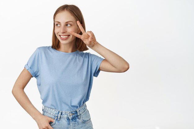 Positive blonde Frau mit kurzen Haaren und weißem Lächeln, zeigt Friedens-V-Zeichen in der Nähe des Gesichts und schaut glücklich beiseite auf Promo-Text, leerer Platz für Logo, stehend gegen weiße Wand.