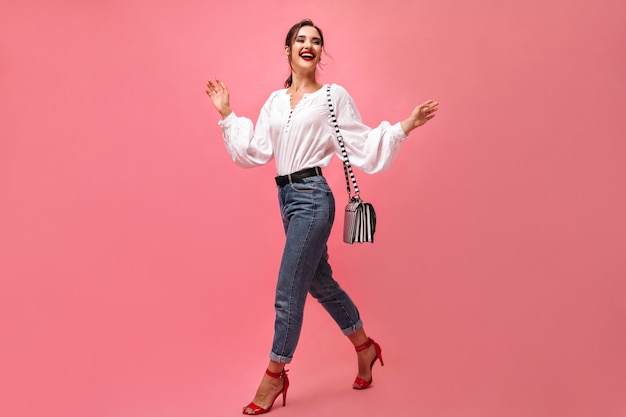 Positive Dame im stilvollen Outfit posiert mit Handtasche. Lächelnde Frau mit rotem Lippenstift und gestreifter Tasche bewegt sich auf lokalisiertem Hintergrund. .