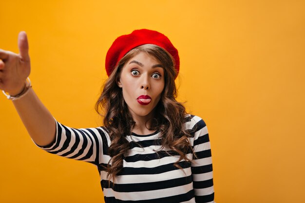 Positive Dame in roter Baskenmütze zeigt Zunge und macht Selfie. Lustige Frau in gestreifter trendiger Bluse und roter Baskenmütze, die auf orange Hintergrund aufwirft.