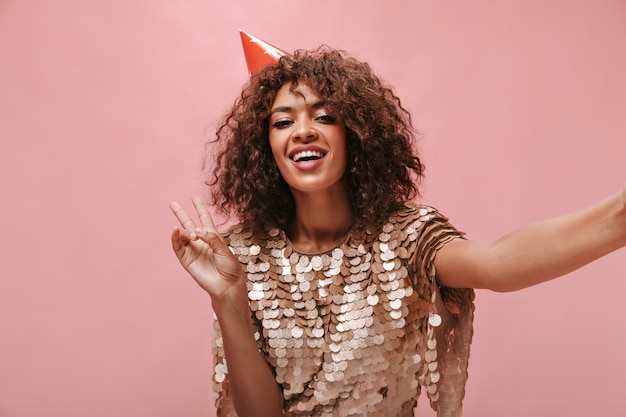 Positive Dame mit welligem Haar in Urlaubsmütze und glänzendem Kleid mit Friedenszeichen und Foto an isolierter rosa Wand..