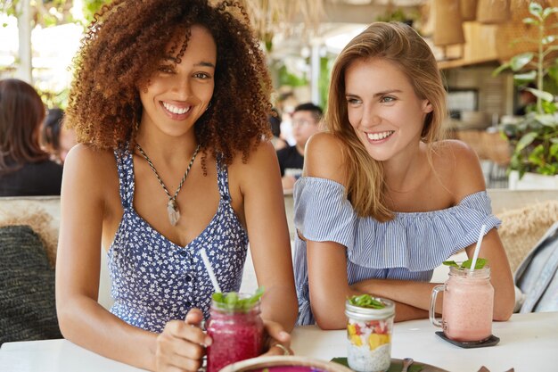 Positive dunkelhäutige afroamerikanische Frau trinkt kalten Smoothie, trifft sich mit der besten Freundin in einem gemütlichen Restaurant, hat fröhliche Gesichtsausdrücke, teilt Neuigkeiten miteinander und plant kommende Ferien.