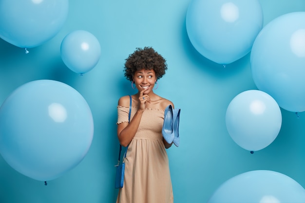 Positive dunkelhäutige Frau in elegantem langem Kleid, wählt blaue Schuhe passend zur Tasche, Kleider, um fabelhaften Blick auf korporative Partei zu haben, steht gegen blaue Wand, fliegende Luftballons herum