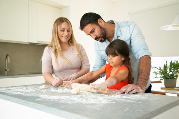 Positive Eltern beobachten, wie Tochter Teig auf Küchenschreibtisch mit Mehl unordentlich rollt. Junges Paar und ihr Mädchen backen Brötchen oder Kuchen zusammen. Familienkochkonzept