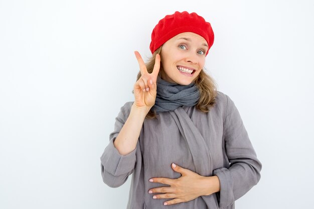 Positive emotionale Französisch Frau zeigt Friedenszeichen und Blick in die Kamera. Verrückte hübsche junge Frau mit zwei Fingern. Handzeichen Konzept