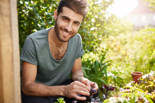 Positive Emotionen, ländlicher Lebensstil. Außenporträt des jungen bärtigen hispanischen Bauern, der mit Zähnen lächelt, in seinem Garten arbeitet, Samen pflanzt, Pflanzen gießt.
