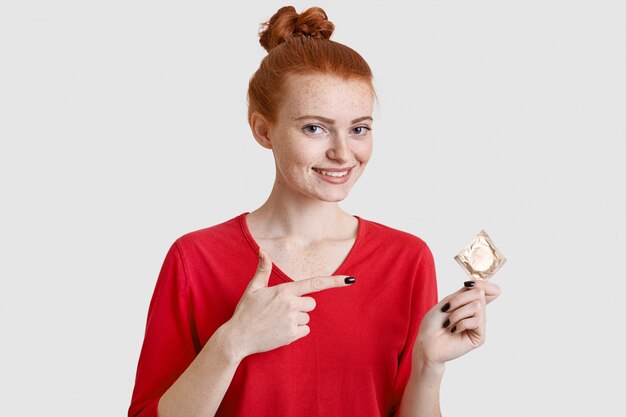 Positive foxy europäische Frau mit sommersprossiger Haut, zeigt auf Kondom, verhindert sich als Sexualleben, trägt rote Kleidung, isoliert über weißer Wand. Menschen, Schwangerschaft und Sicherheitskonzept