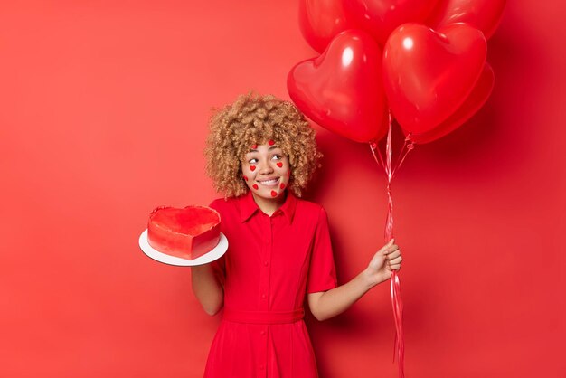 Positive Frau hat lockiges blondes Haar, trägt ein festliches Kleid, hält einen Haufen Herzballons und süßen Kuchen bereitet sich auf den Urlaub vor, der über leuchtend rotem Hintergrund isoliert ist Menschen- und Feierkonzept