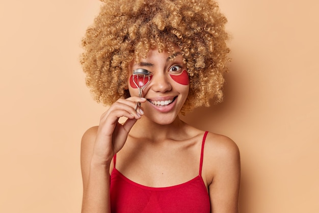 Positive Frau hat lockiges, buschiges Haar, verwendet Wimpernzange für Make-up, trägt Hydrogel-Patches unter den Augen auf, die in einem lässigen roten T-Shirt gekleidet sind