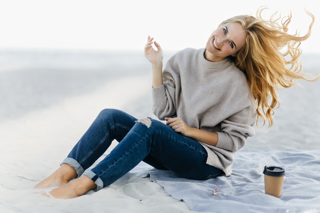 Positive Frau im weichen Pullover, der am Strand herumalbert. Außenporträt des charmanten weiblichen Modells, das im Sand mit Tasse Tee sitzt.