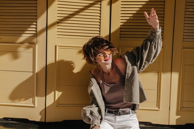 Positive Frau in Jeansjacke und Unterhemd blickt auf die Sonne auf hellem Hintergrund Brünettes Mädchen in leichter Hose und Sonnenbrille posiert auf gelbem Hintergrund