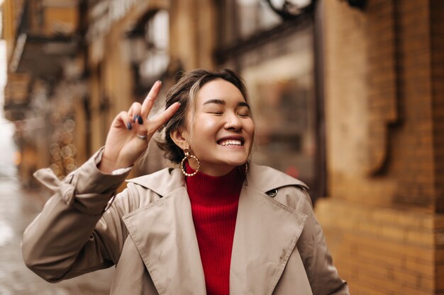 Positive Frau in massiven Ohrringen, die mit geschlossenen Augen gegen Wand des Gebäudes lachen