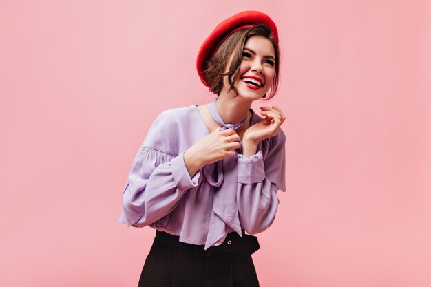 Positive Frau in roter Baskenmütze und lila Bluse lacht auf rosa Hintergrund.