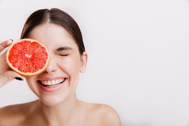 Positive Frau mit einem Lächeln schloss die Augen. Mädchen mit gesunder Haut wirft mit Grapefruit auf isolierter Wand auf.