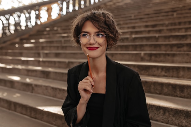 Positive Frau mit kurzen Haaren und hellen Lippen in den Gläsern, die draußen lächeln. Trendy Dame in der schwarzen Kleidung, die auf Treppen aufwirft.