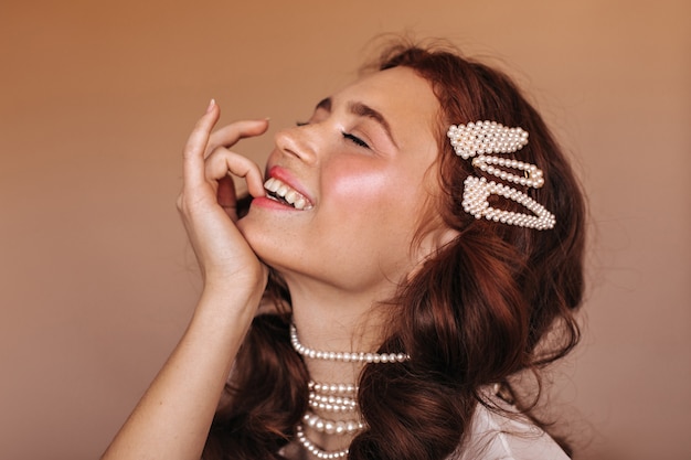Positive Frau mit lockigem Haar lacht und beißt sich auf den Finger. Porträt der Frau mit weißen Haarnadeln und Perlenkette.