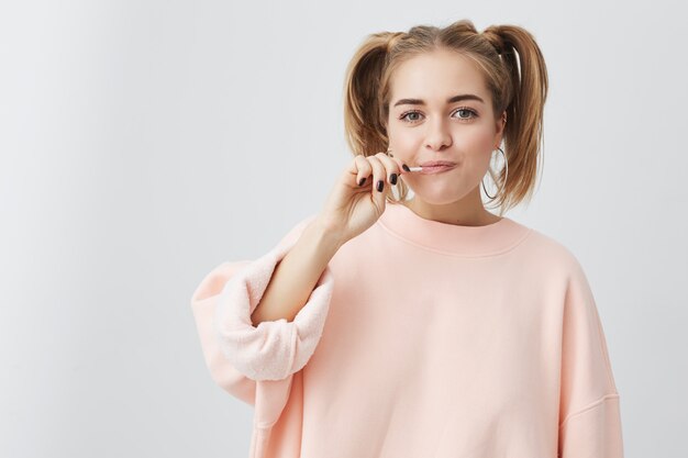 Positive Frau mit lustigem Blick, verspielt, losen rosa Pullover tragend, mit Lutscher im Mund, posierend. Glückliches junges Mädchen mit zwei Pferdeschwänzen, die positive Emotionen zeigen.