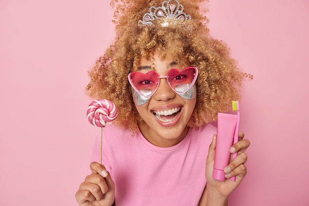 Positive frau mit natürlichem lockigem haar lächelt zahnig hält lollipop-zahnbürste und zahnpasta-tube trägt silberne schönheitsflecken unter den augen, um die haut mit feuchtigkeit zu versorgen, trägt herz-sonnenbrillen-t-shirt