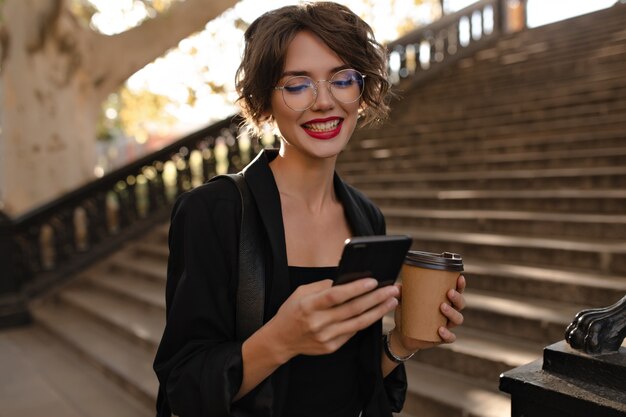 Positive Frau mit roten Lippen im schwarzen Outfit, das mit Telefon und Tasse Kaffee aufwirft. Lockige Frau in den Gläsern, die draußen lächeln.