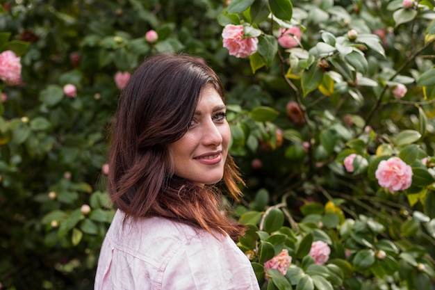 Kostenloses Foto positive frau nahe den rosa blumen, die auf grünen zweigen des busches wachsen