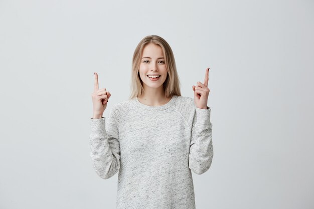 Positive freudige blonde Frau, die mit Zeigefingern auf Kopienraum zeigt.