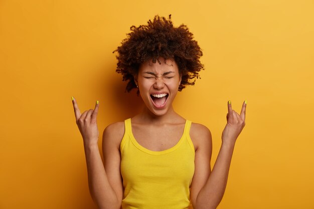 Positive freudige Frau macht Heavy Metal Zeichen, hat Spaß am Musik-Rocking-Festival, ruft laut aus, schließt die Augen, gestikuliert aktiv, in Freizeitkleidung gekleidet, isoliert über gelber Wand. Rock'n'Roll Baby