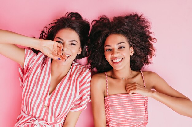 Positive Freundinnen, die sich hinlegen und Ruhe im Zimmer des rosa Mädchens genießen. Porträt gebräunter Brünetten in gestreiften Sommerkleidern.