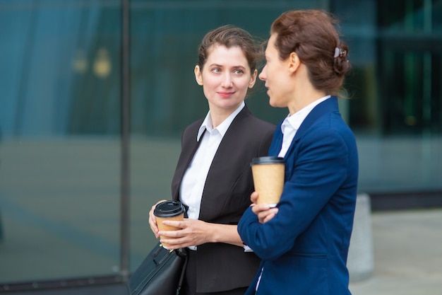 Positive Freundinnen im Büro mit Kaffeetassen zum Mitnehmen, die zusammen im Freien spazieren gehen, über Projekte sprechen oder sich unterhalten. Mittlerer Schuss. Arbeitspausen-Konzept