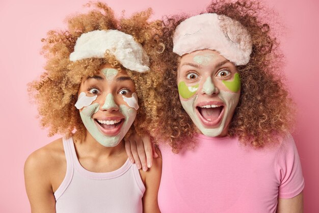 Positive, fröhliche Frauen sehen mit verblüfften Ausdrücken aus. Sie werden Schönheitsverfahren unterzogen, wenden Patches und Hautpflegemasken an, tragen Schlafmasken, lässige T-Shirts, die über rosafarbenem Hintergrund isoliert sind. Wellness-Konzept