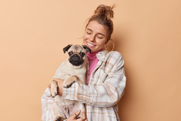 Positive, fürsorgliche junge Europäerin umarmt ihren Pug-Hund demonstriert eine romantische Beziehung zwischen Mensch und Haustier, verbringt gemeinsam Freizeit, posiert vor braunem Hintergrund, spielt und hat Spaß