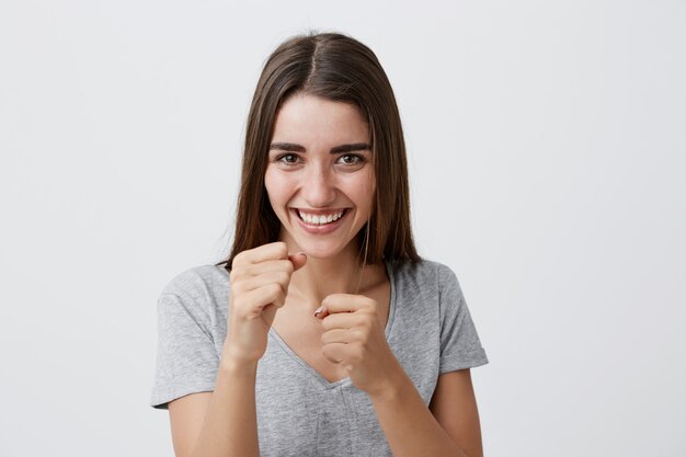 Positive Gefühle. Glückliches junges hübsches freudiges Mädchen mit langen braunen Haaren im lässigen einfachen T-Shirt lachend, Händchen haltend vor ihr in Kampfhaltung, mit fröhlichem Gesichtsausdruck.