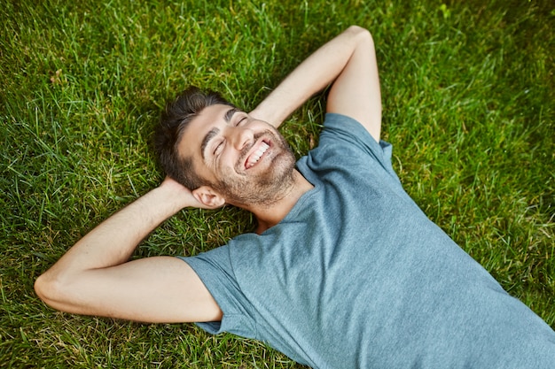 Kostenloses Foto positive gefühle. junger schöner bärtiger kaukasischer mann im blauen t-shirt liegend auf gras lächelnd mit zähnen, lachend, entspannend draußen im sommermorgen mit glücklichem gesichtsausdruck.