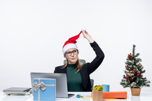 Positive Geschäftsfrau, die mit ihrem Weihnachtsmannhut spielt, der an einem Tisch mit einem Weihnachtsbaum und einem Geschenk darauf sitzt und sich auf etwas auf weißem Hintergrund konzentriert